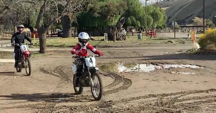A student riding a dirt bike around a puddle on the training course with an instructor following behind